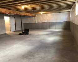 Unfinished basement with concrete floor, exposed wooden beams, and partial walls covered in plastic sheeting. Some tools on the ground.