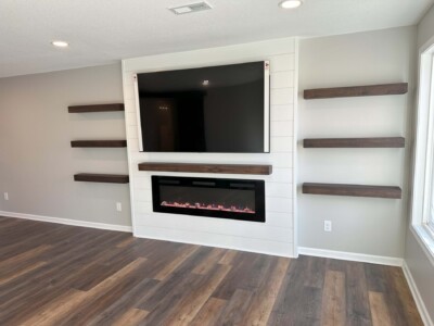 Living room with a wall-mounted TV above an electric fireplace, flanked by five floating wooden shelves on a gray wall.