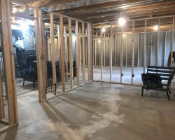 A basement under construction with wooden framing, exposed beams, and concrete floor, illuminated by hanging light bulbs.
