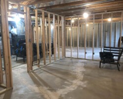 A basement under construction with wooden framing, exposed beams, and concrete floor, illuminated by hanging light bulbs.