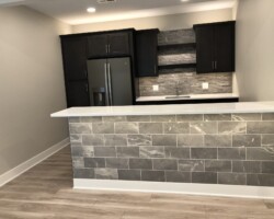 Modern kitchen with dark cabinets, stone backsplash, gray tiled island, and stainless steel refrigerator.